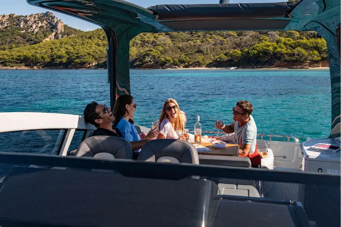 Four friends enjoying a meal on a boat, laughing with drinks, surrounded by blue water and green hills in the background.